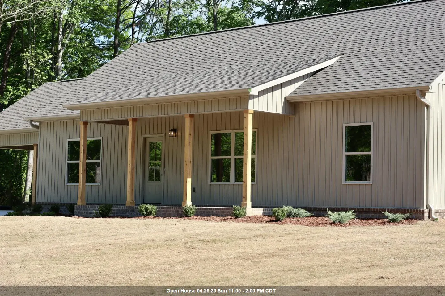 Beige house with a gray roof, wooden porch columns, and a green lawn. Trees are visible in the background.