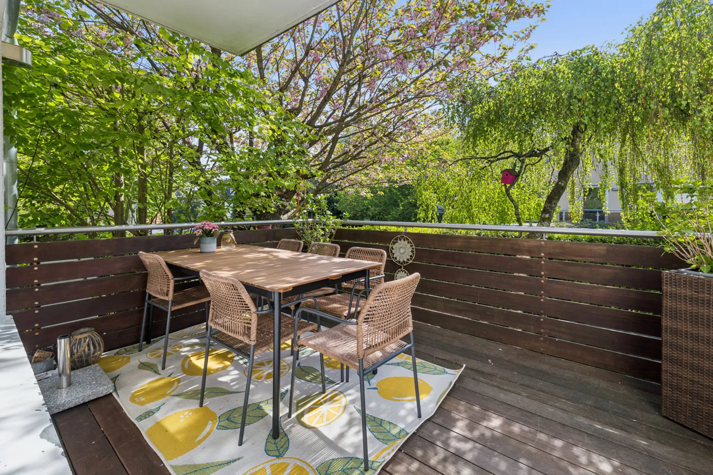 Outdoor patio with a wooden table, wicker chairs, and a lemon-patterned rug, surrounded by lush green trees.