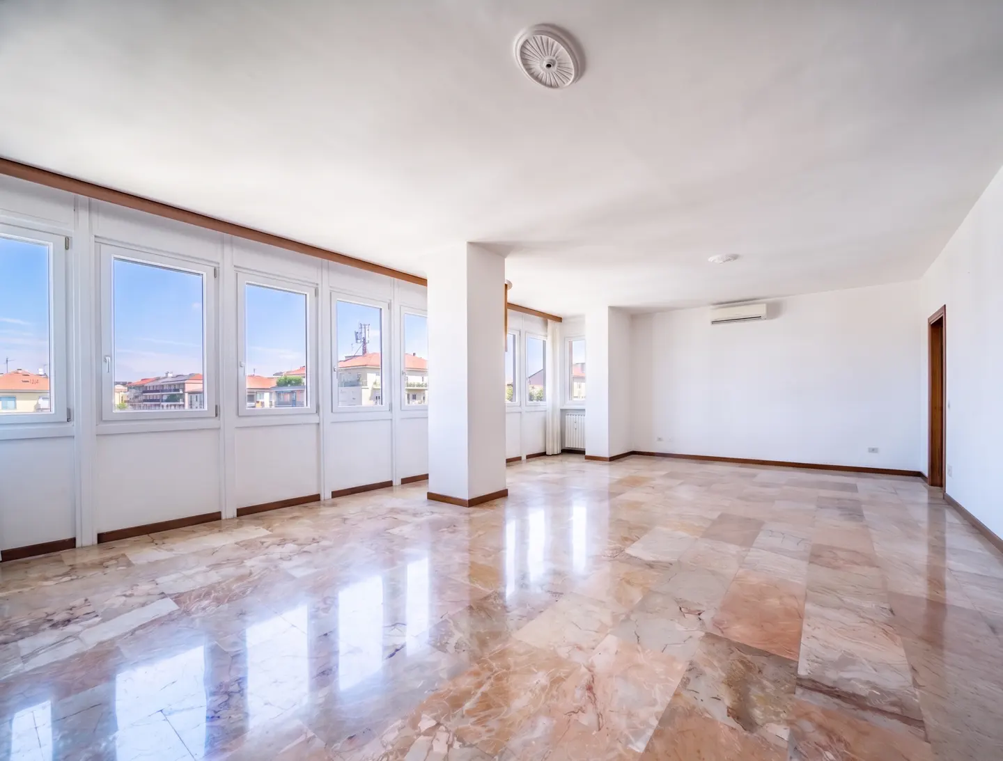 Bright, empty room with marble floors, white walls, and many windows showing a city view.