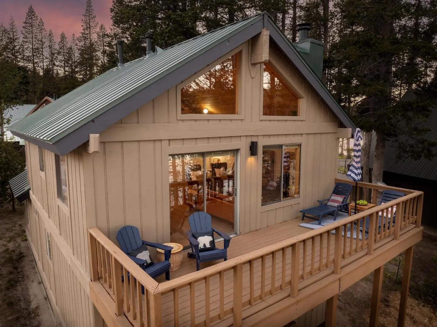 Tan cabin with a green metal roof and a wooden deck featuring blue Adirondack chairs, set against a forest at sunset.