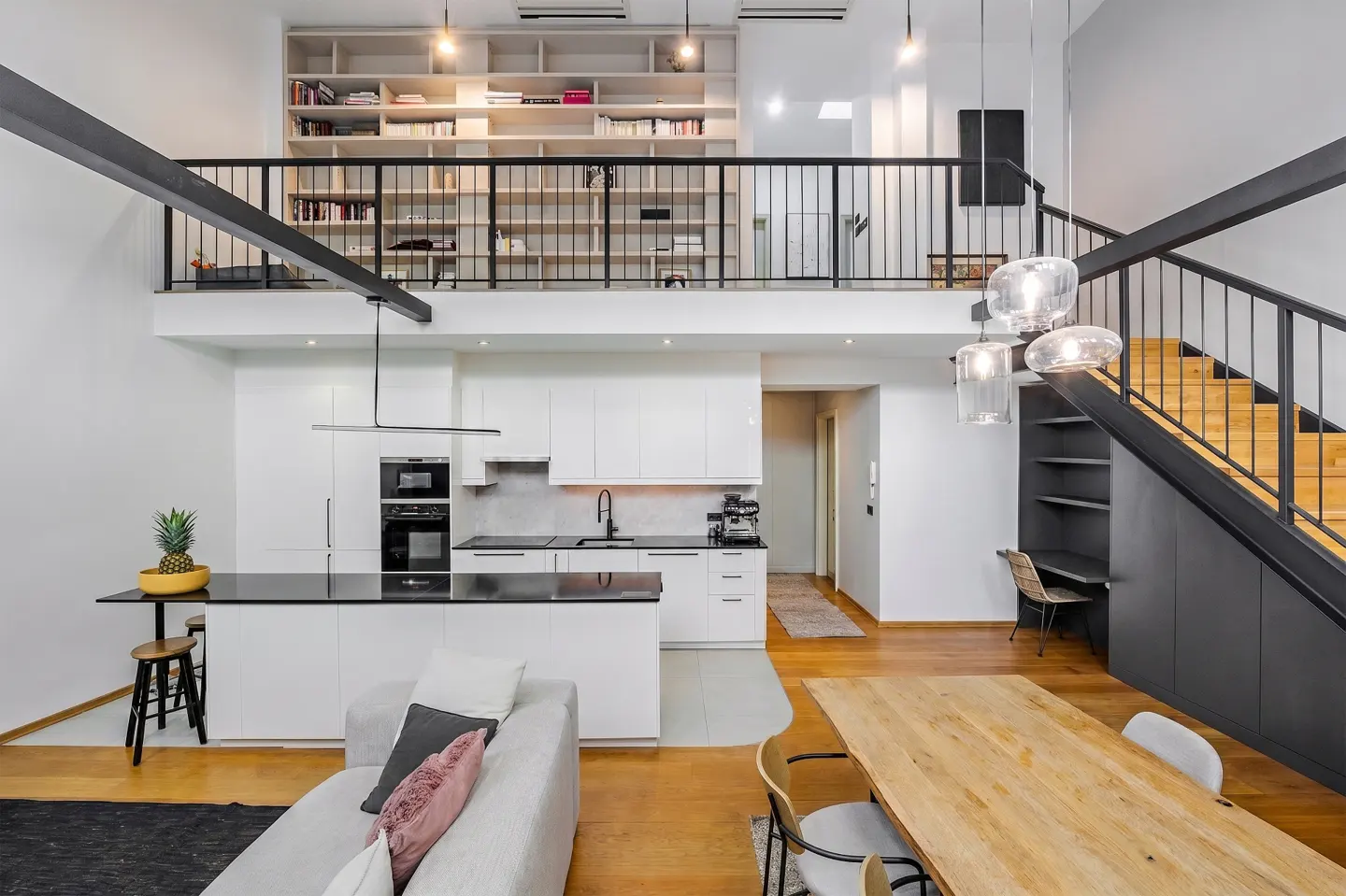A modern, open-concept loft apartment with white kitchen, wood floors, and black staircase. A bookshelf lines the upper level.