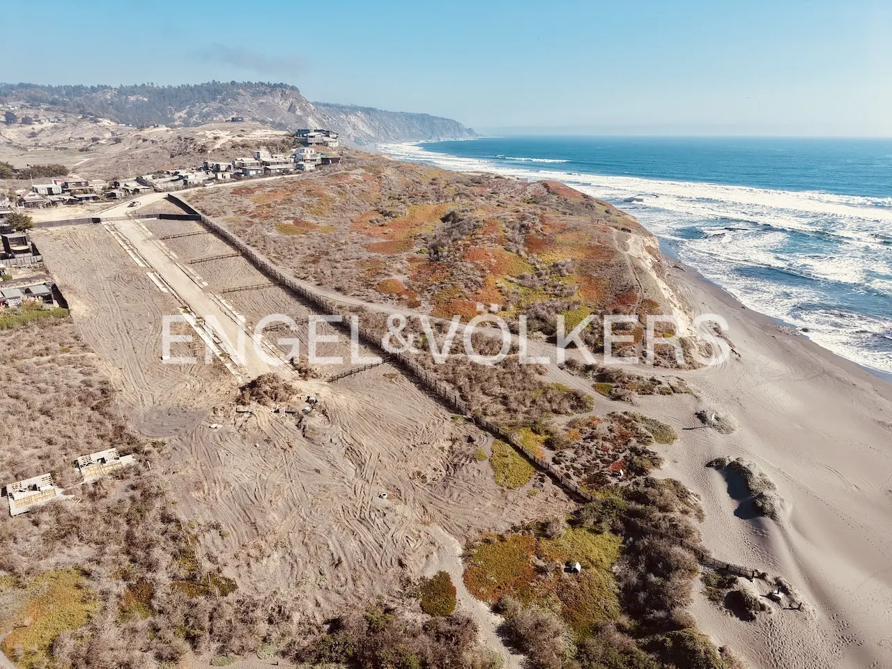 Aerial view of a sandy beach with waves, dunes, and vegetation under a blue sky. The Engel & Völkers logo is superimposed on the image.