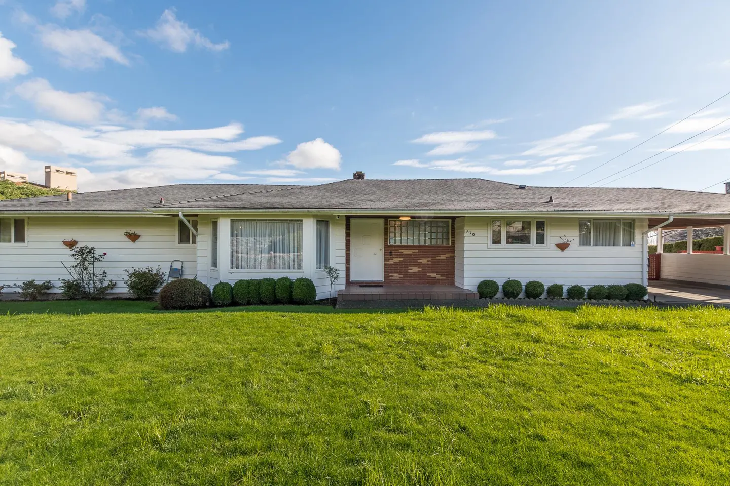 A white, single-story house with a gray roof and green lawn under a blue sky. A carport is visible on the right side.