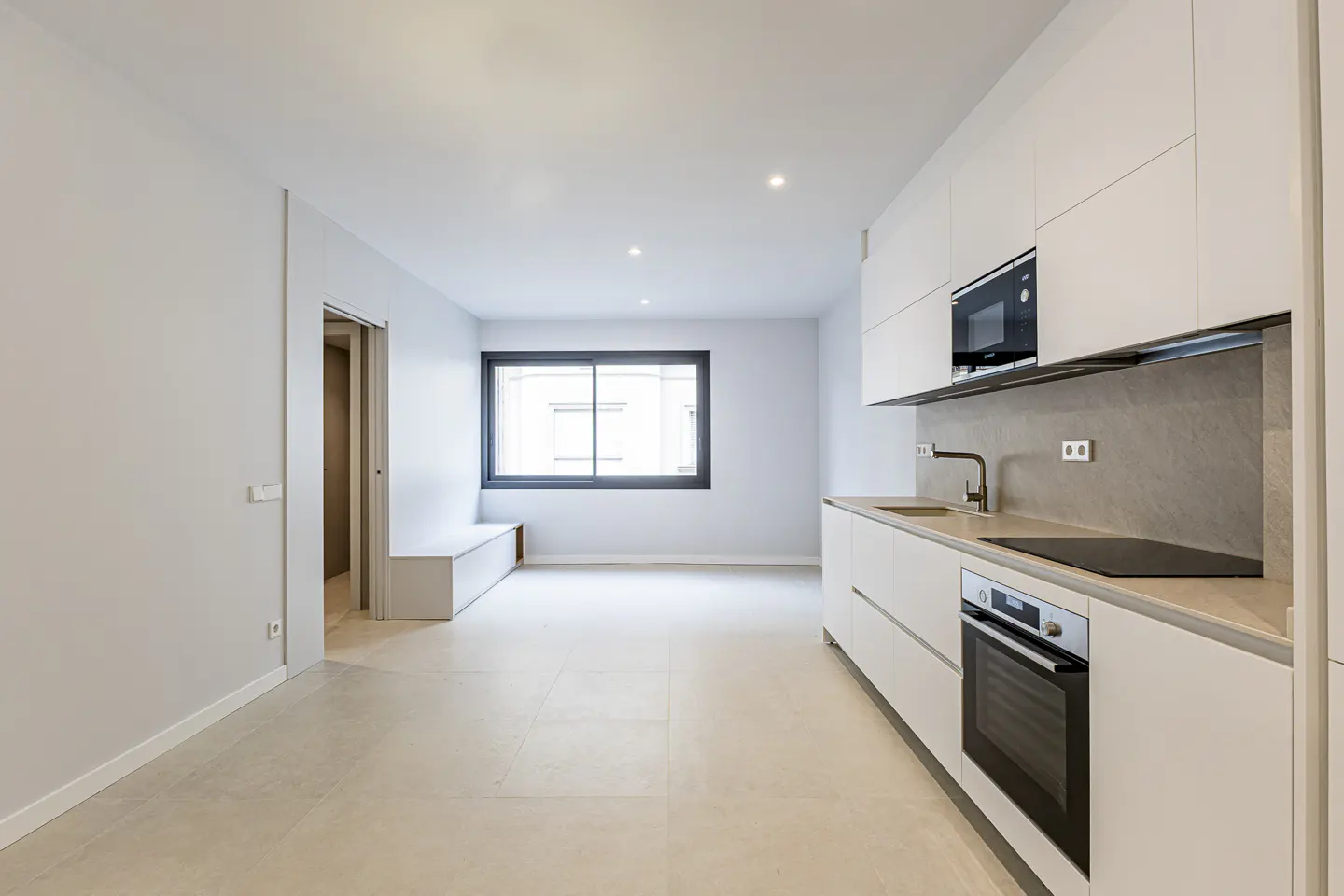 Modern minimalist kitchen with white cabinets, built-in appliances, and light tile floors in an open-plan apartment.