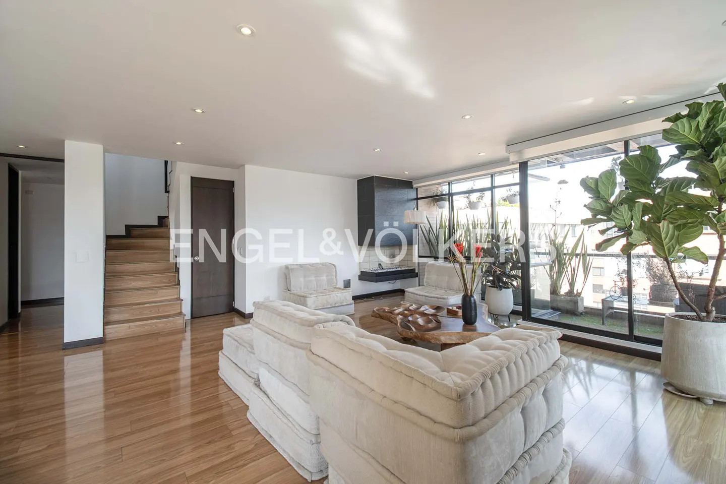 Bright living room with hardwood floors, white modular sofa, and floor-to-ceiling windows with plants. Stairs lead to an upper level.