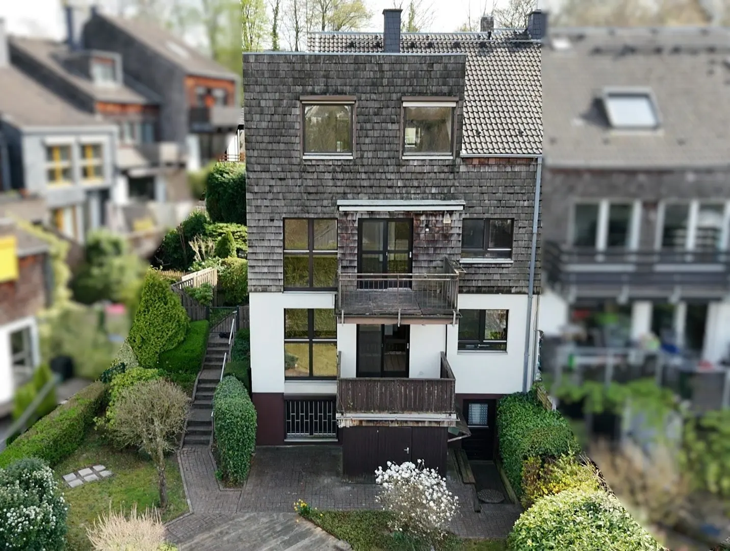 A three-story house with gray shingles and white siding, surrounded by greenery. Balconies are visible on the second and third floors.