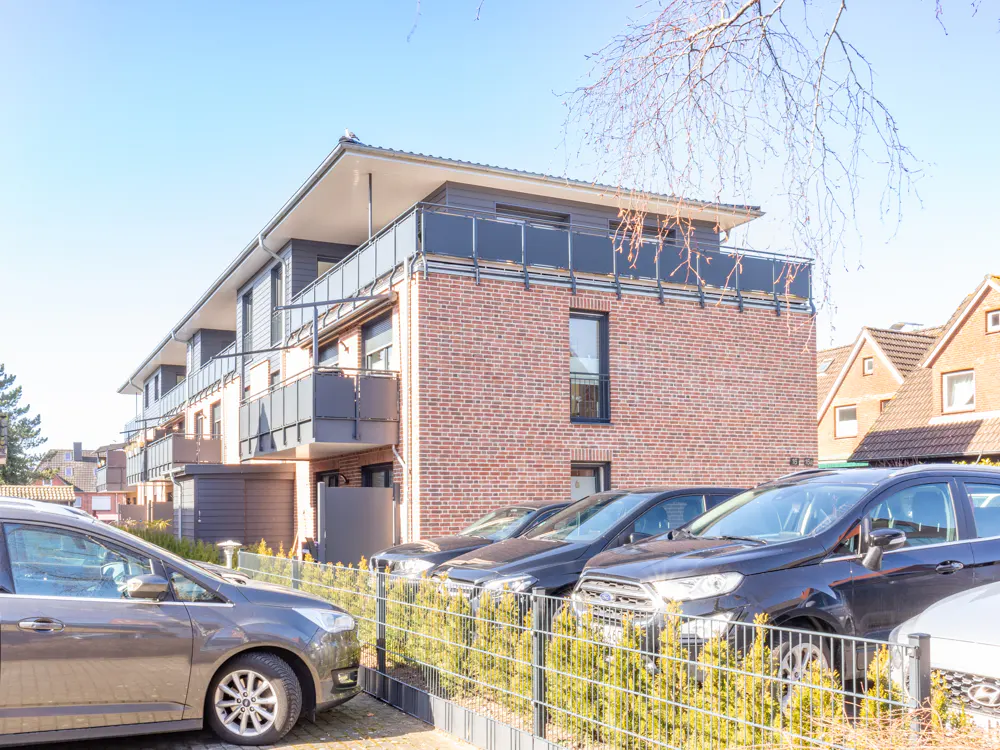 Modern brick apartment building with balconies and parked cars in front on a sunny day.