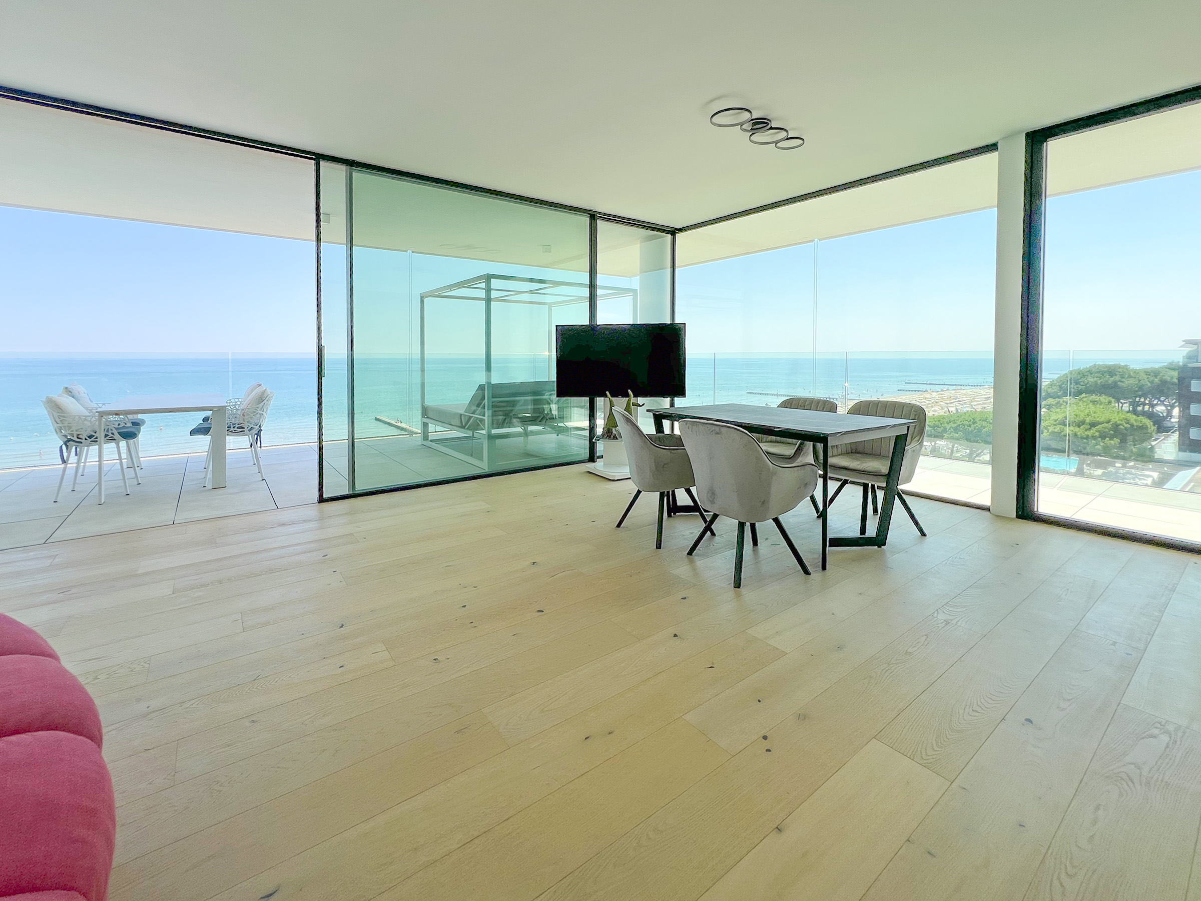 Modern living room with light wood floors, dining table, and floor-to-ceiling glass walls overlooking the ocean.