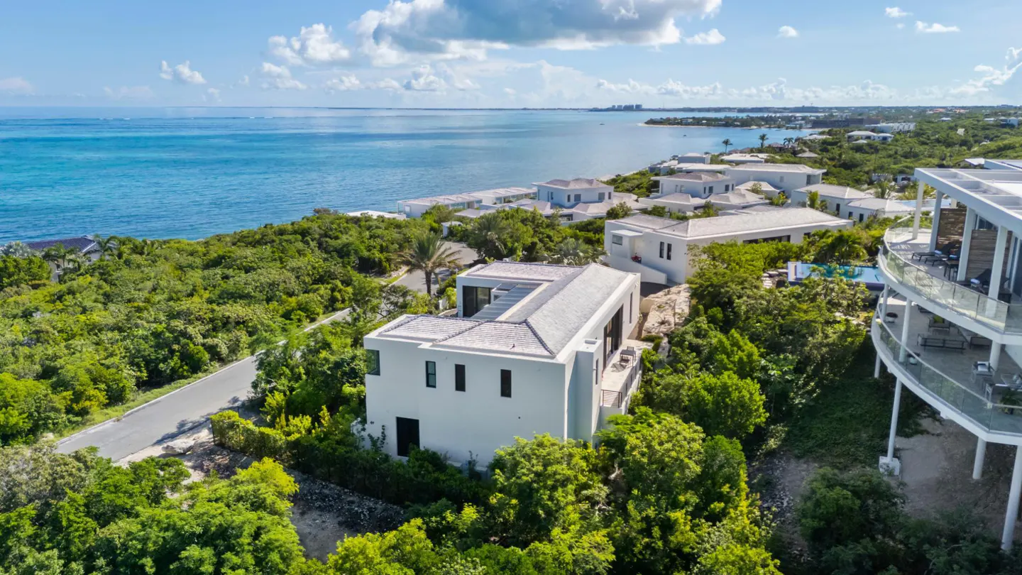 Aerial view of modern white houses nestled in lush green trees near a turquoise ocean under a partly cloudy sky.