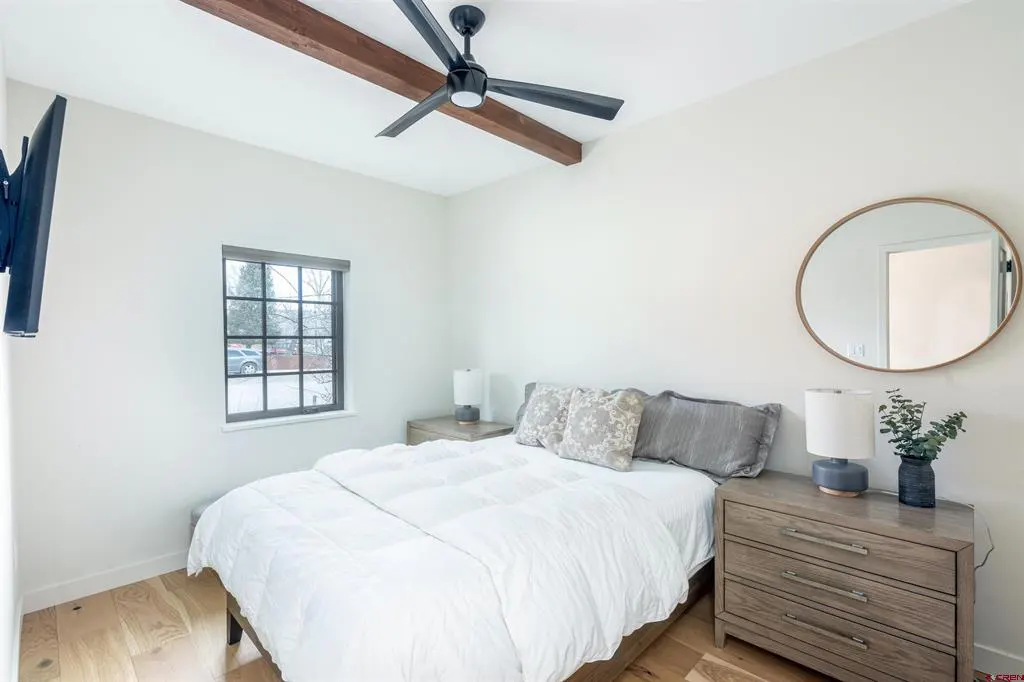 A bright bedroom with a white comforter on the bed, a ceiling fan, and a round mirror.