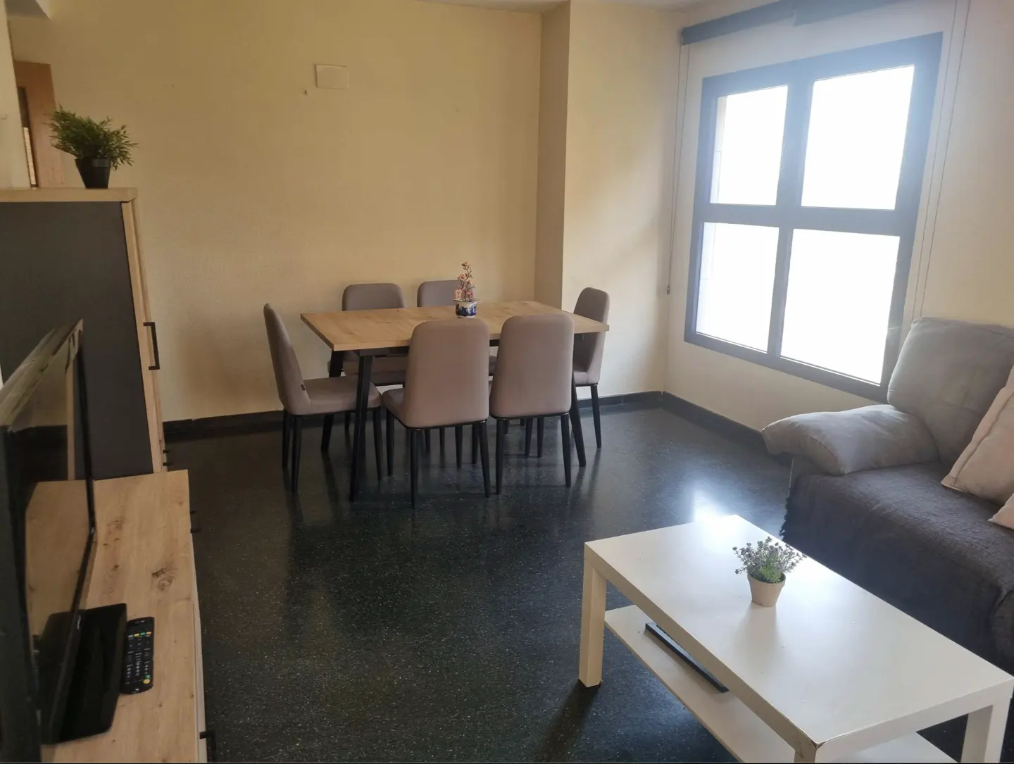 Living room with a wooden dining table, six beige chairs, a grey sofa, white coffee table, and large window.