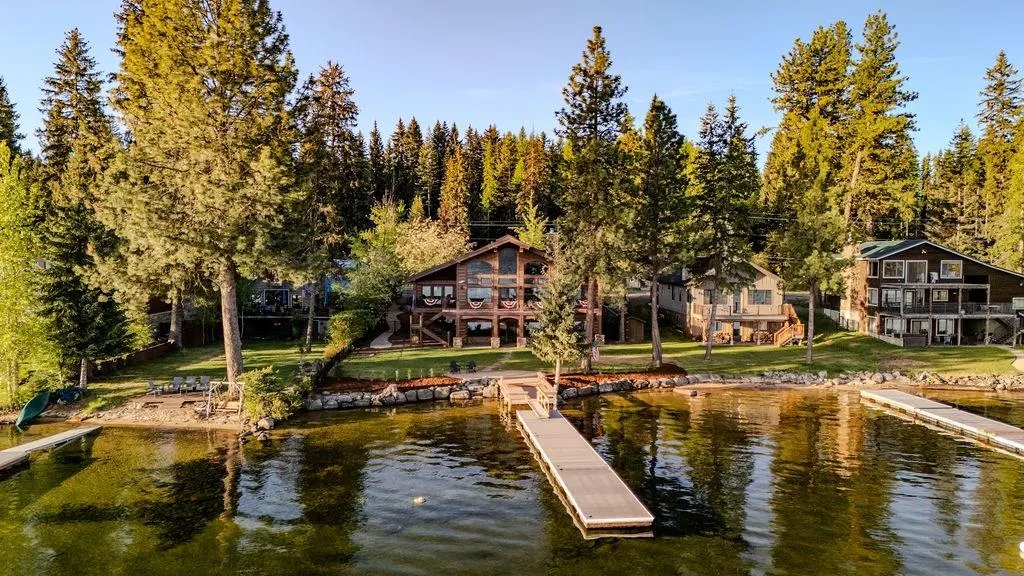 Lakefront homes with docks. A large log cabin is centered, surrounded by tall green trees and calm water.