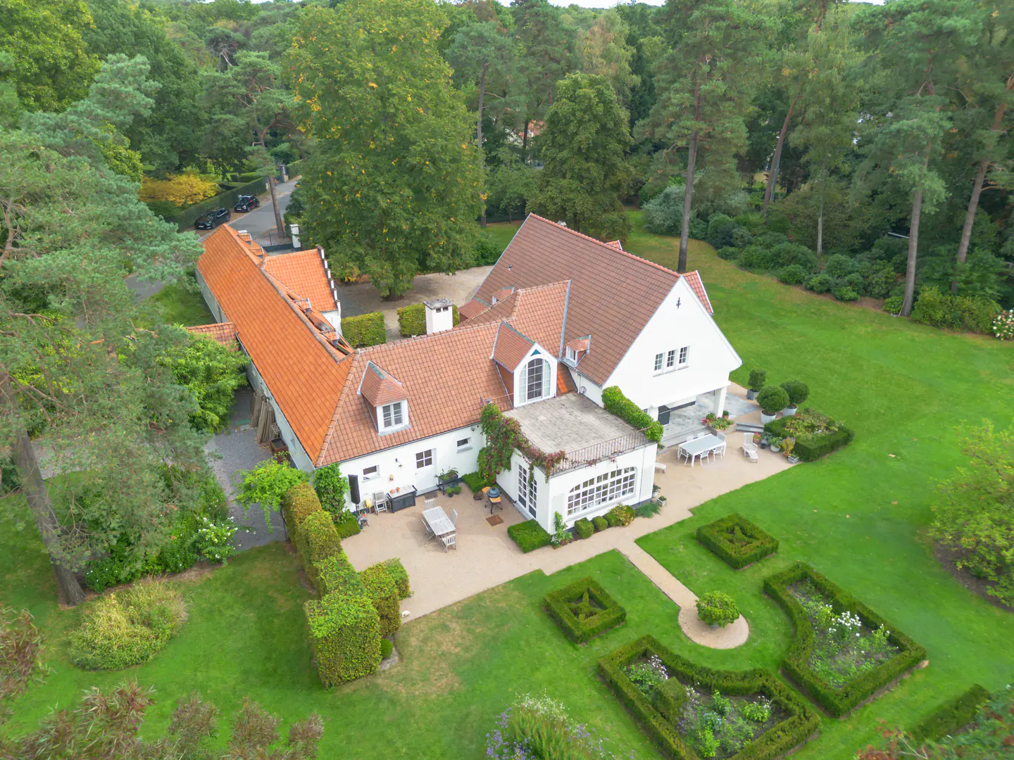 Aerial view of a large white house with a red tile roof, surrounded by green lawns, trees, and a formal garden.