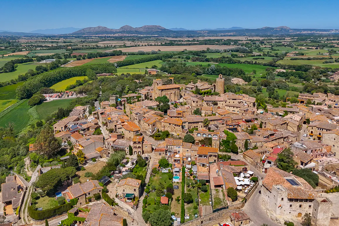 Aerial view of Pals, Spain, a medieval town with stone buildings and red tile roofs, surrounded by green fields and distant mountains under a blue sky.