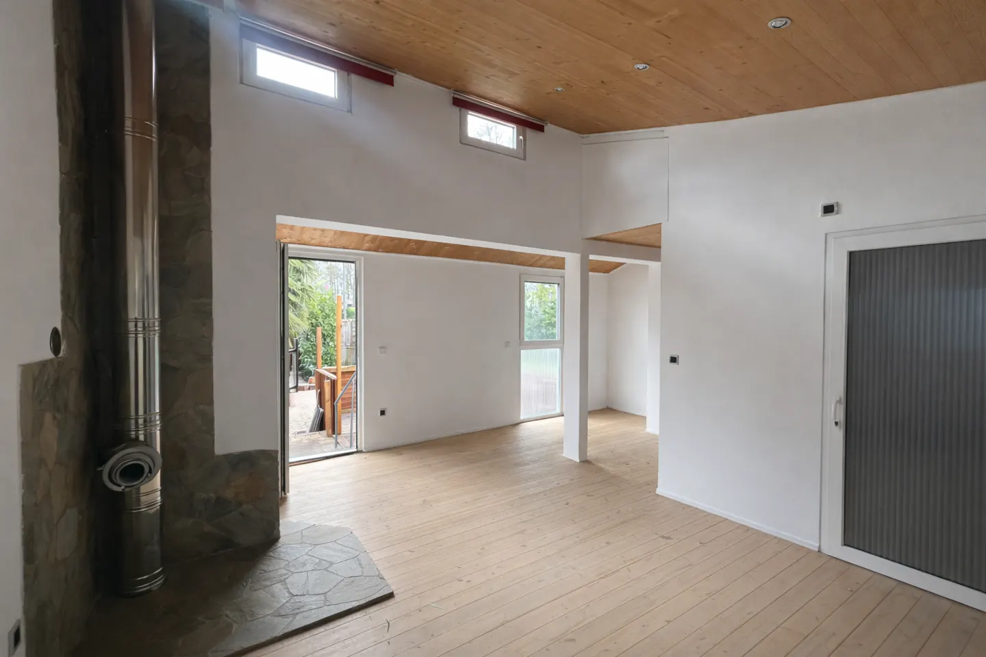 Bright, empty room with wood floors, white walls, and wood ceiling. A stone fireplace and sliding glass door are visible.