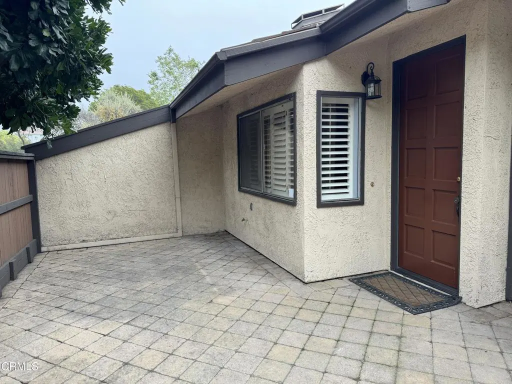 Exterior view of a beige stucco house with a brown door, window with white shutters, and a gray brick patio.