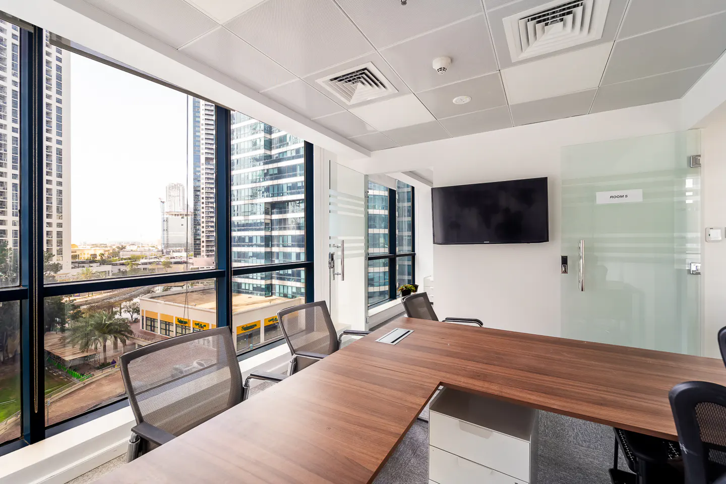 Bright, modern office space with a large window view of city buildings. A wood conference table and black chairs are centered. A TV is mounted on the wall.