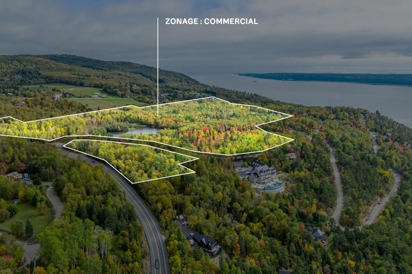 Aerial view of commercial zoned land with white outline, surrounded by colorful autumn trees, a lake, and a distant body of water.
