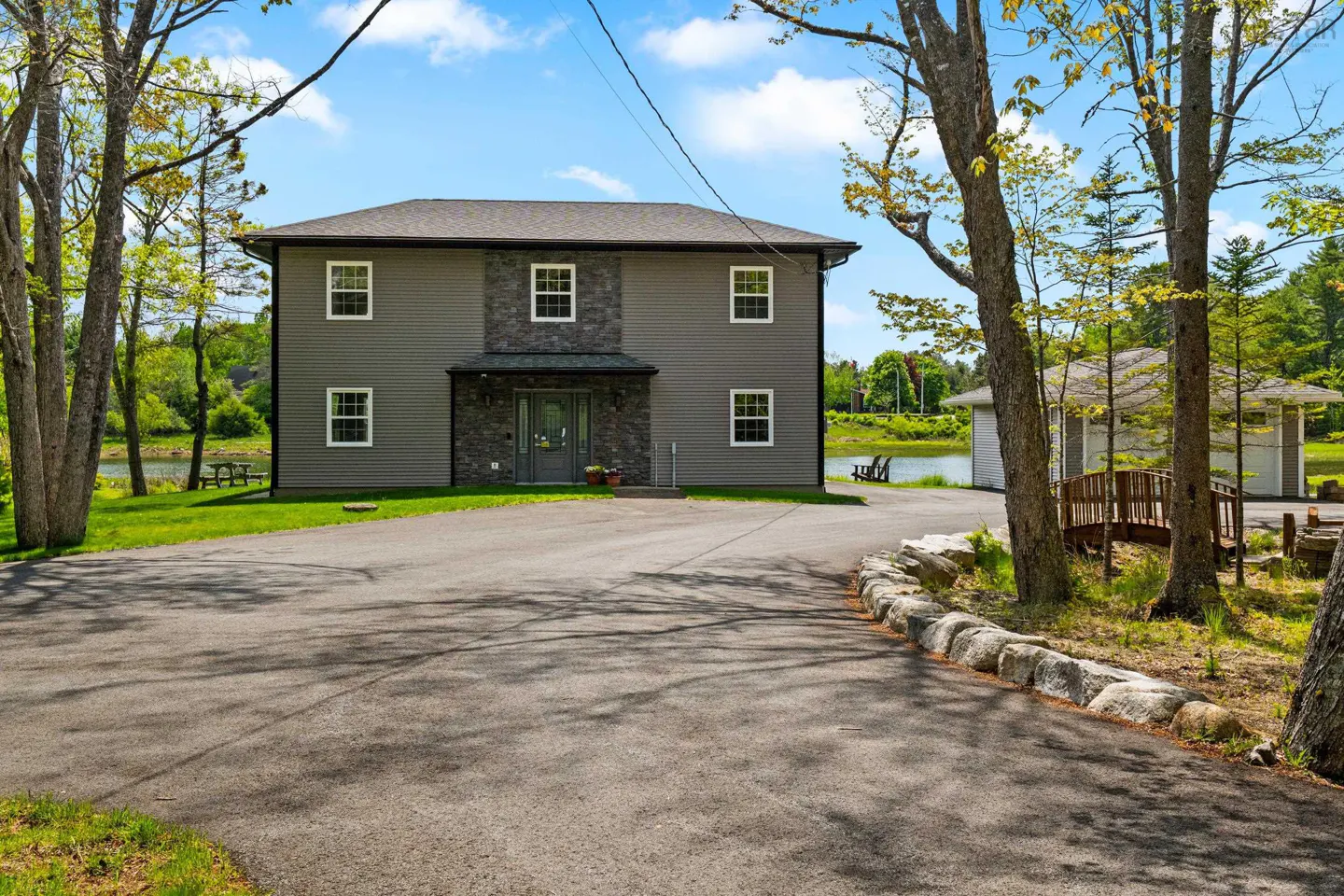 Two-story gray house with stone accents, a paved driveway, and a small wooden bridge near a pond on a sunny day.