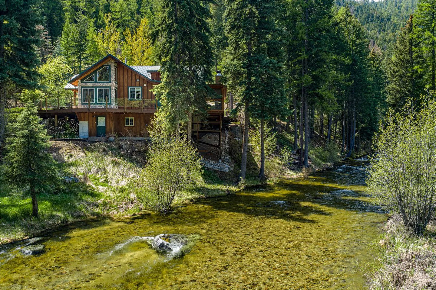 A wood and stone house sits on a hill overlooking a clear, shallow river surrounded by green trees.