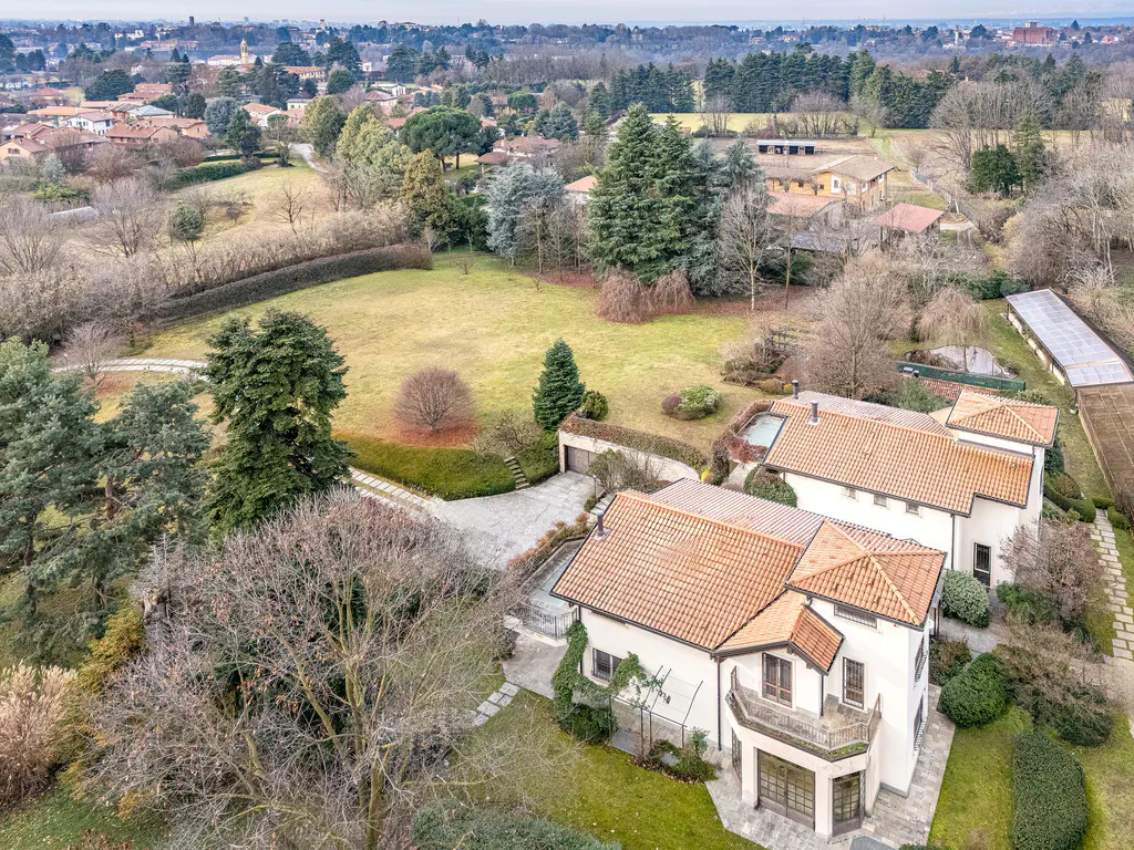 Aerial view of a large, white house with a red tile roof, surrounded by green lawns and trees. A driveway leads to a garage.