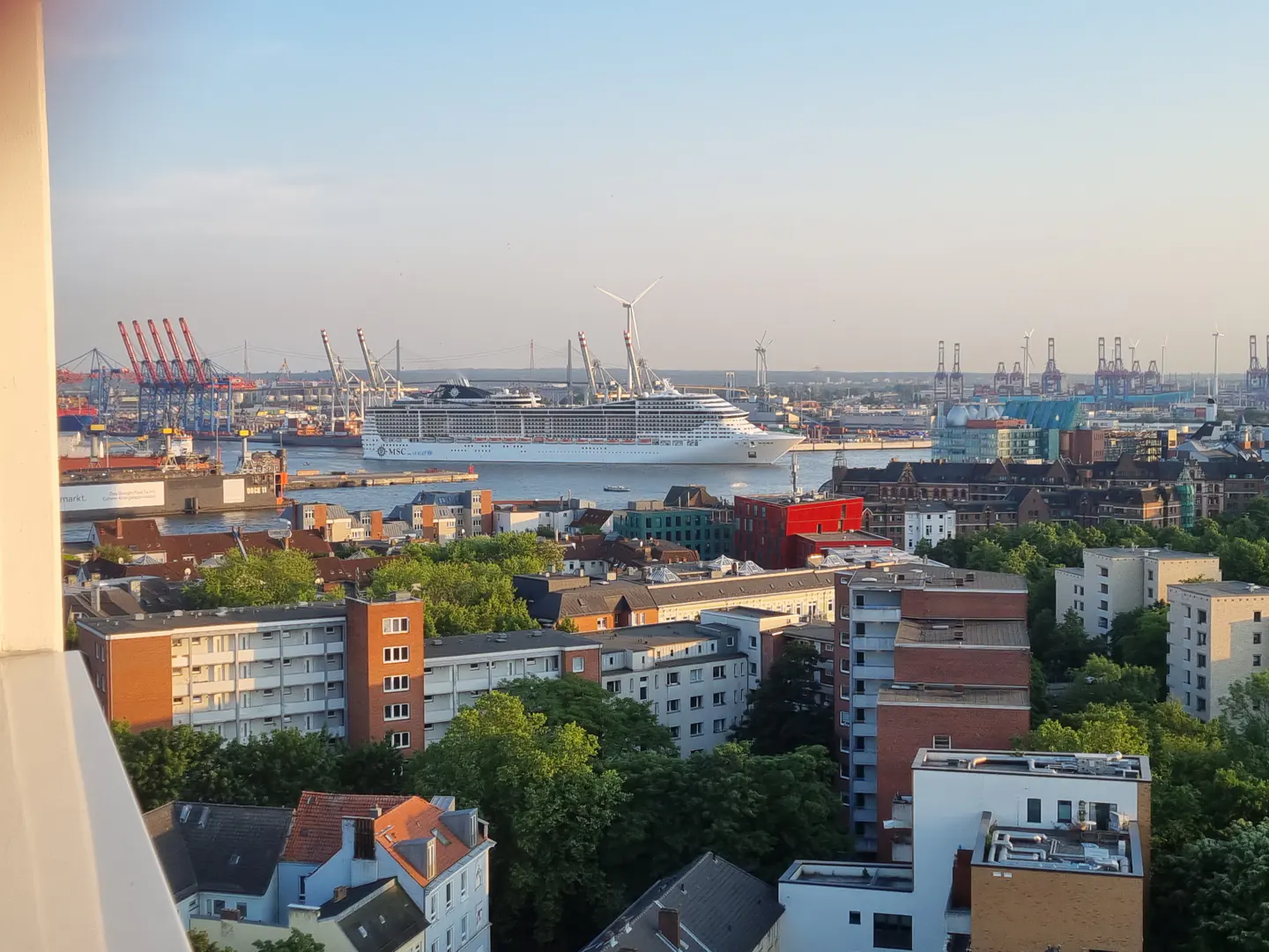 Cityscape view with a cruise ship in the harbor, surrounded by buildings, cranes, and wind turbines under a blue sky.