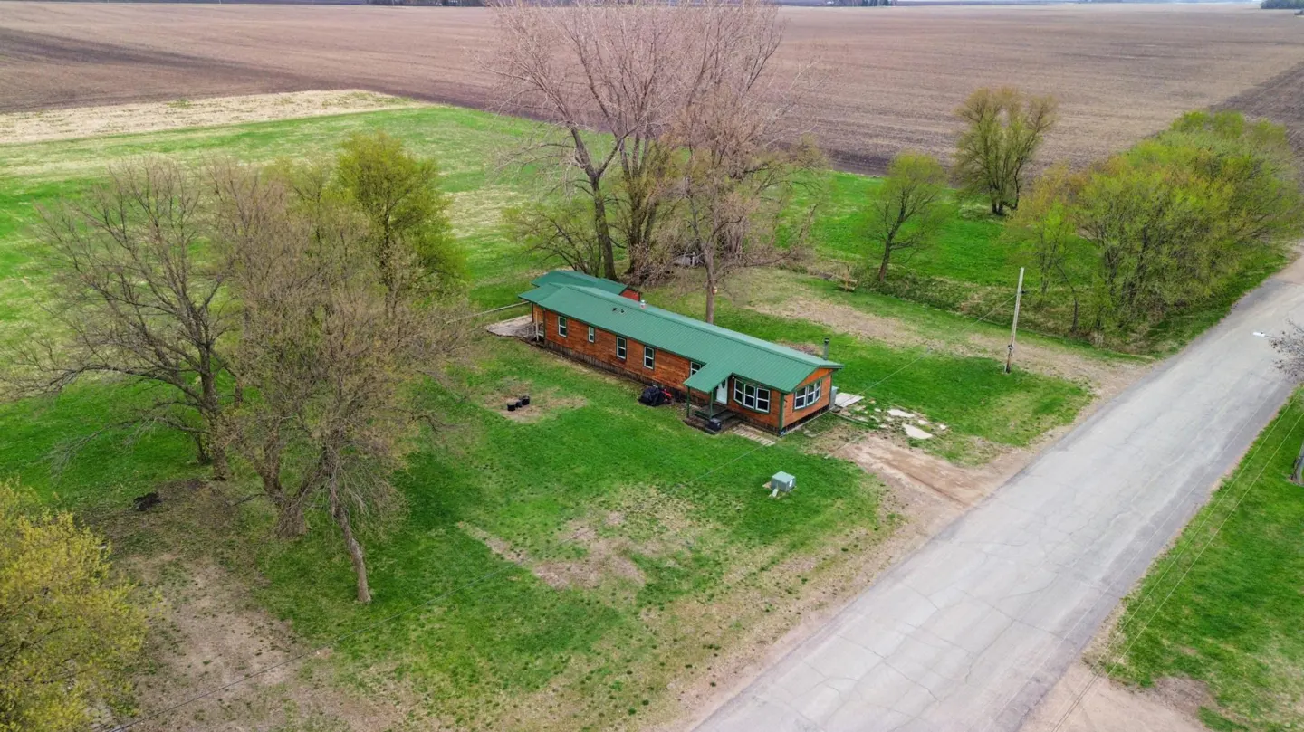 Aerial view of a wood-sided home with a green roof, surrounded by a green lawn and trees, next to a road and farm fields.