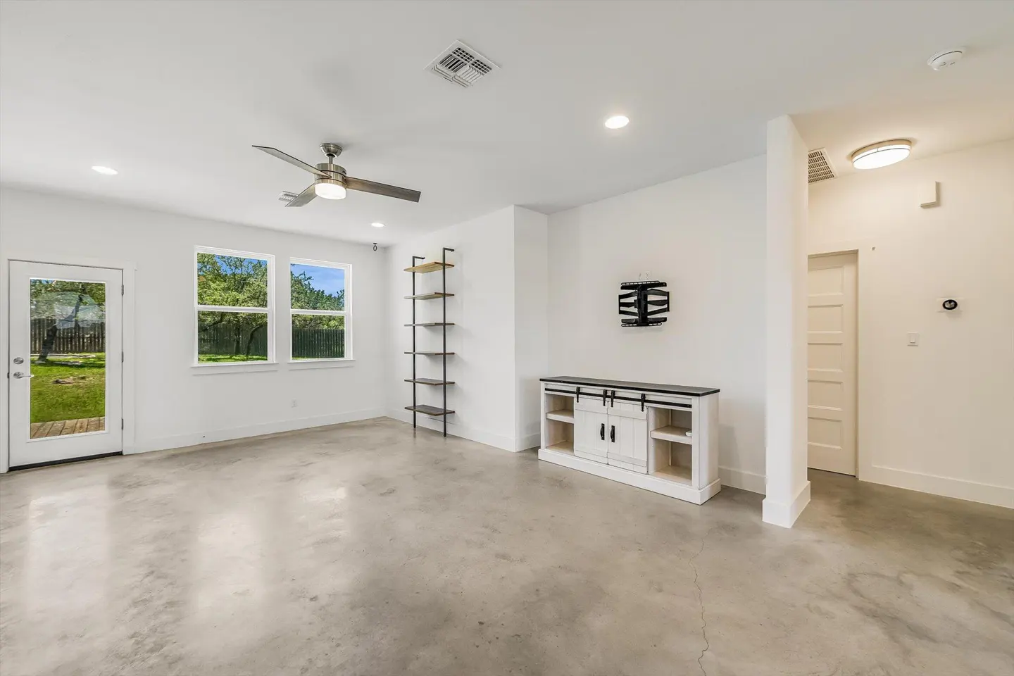 Bright, modern living room with white walls, concrete floors, and a ceiling fan. A sliding door cabinet and open shelves add storage. Windows view the green yard.
