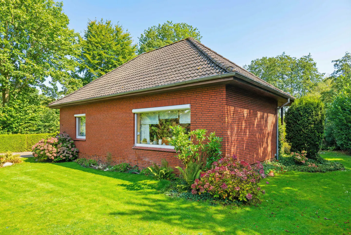Exterior view of a red brick house with a brown tiled roof, green lawn, and lush trees. Windows have white frames.