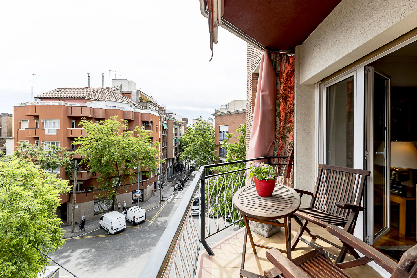 Balcony view with wooden chairs, table, and flower pot overlooking a city street with buildings and trees.