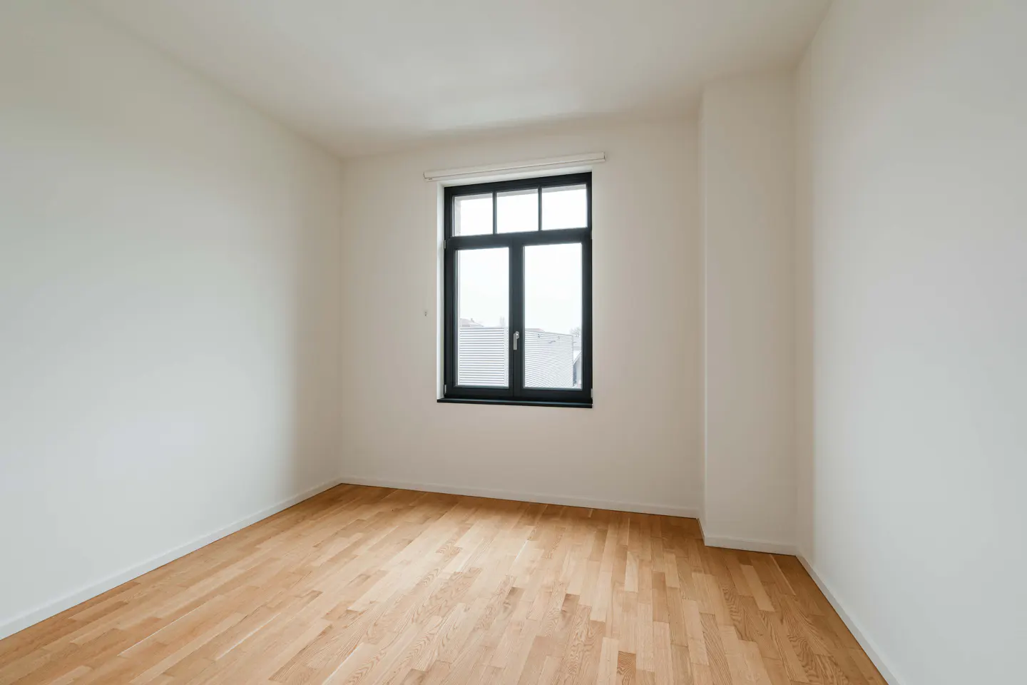 Bright, empty room with light wood floors, white walls, and a black-framed window showing a cityscape.