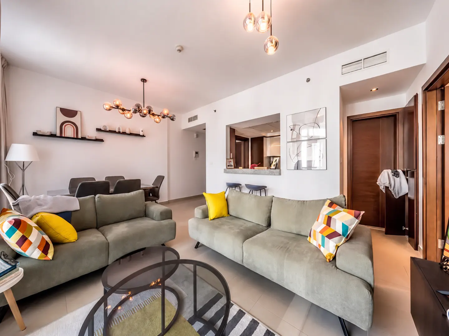 Bright living room with two olive green sofas, colorful pillows, and glass coffee tables. Dining area and kitchen visible in the background.