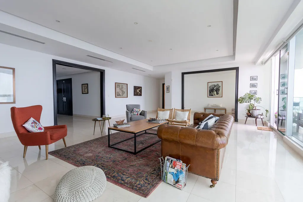 Bright living room with a brown leather sofa, red chair, and a patterned rug on a white tile floor. Artwork adorns the white walls.