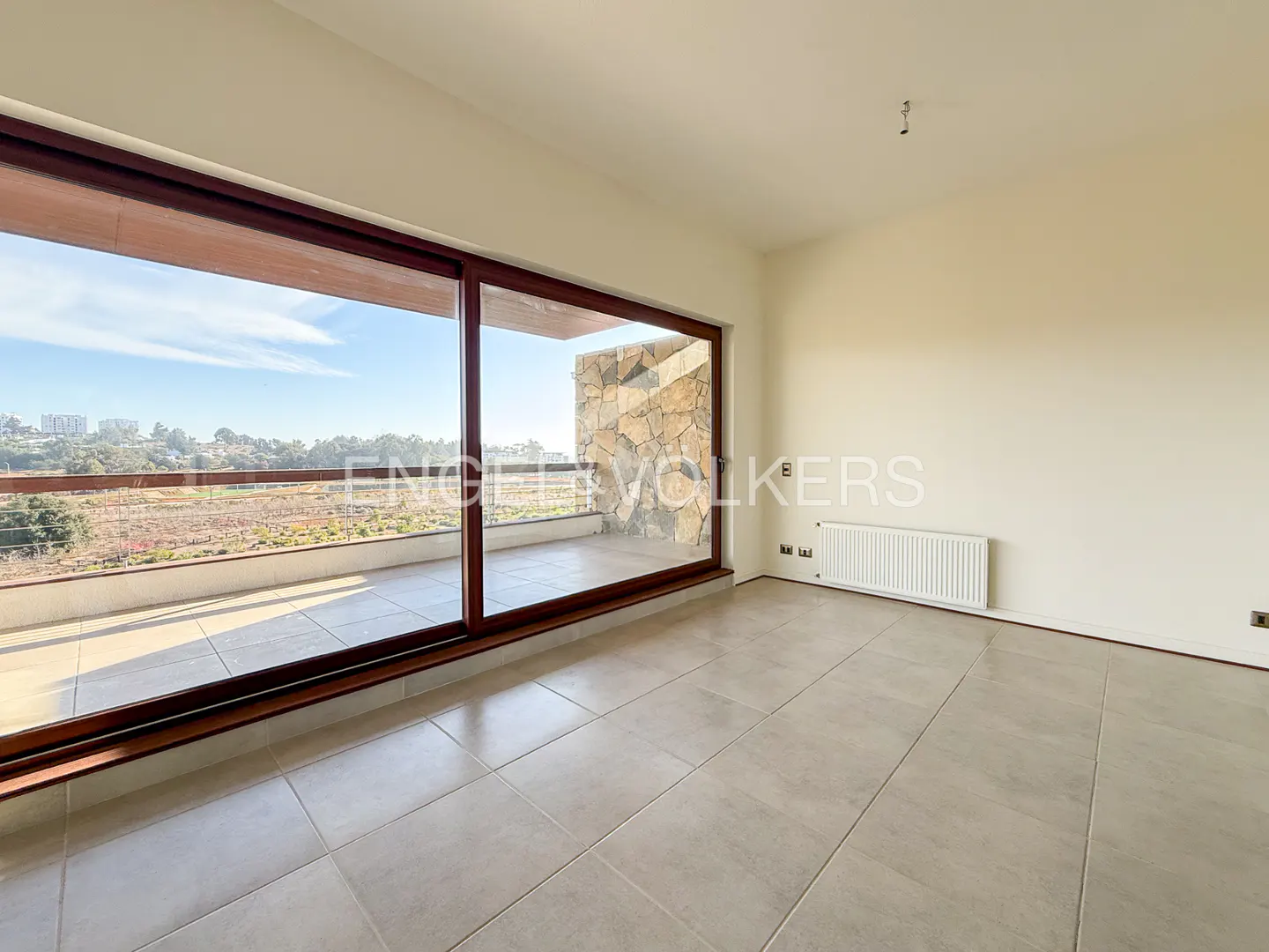 An empty room with beige tile floors and walls, a radiator, and a large window with a brown frame overlooking a balcony and landscape.
