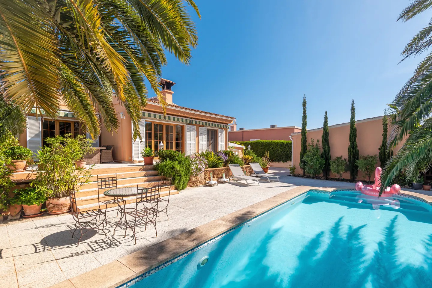 Sunny backyard with a swimming pool, patio furniture, palm trees, and a peach-colored house under a clear blue sky.