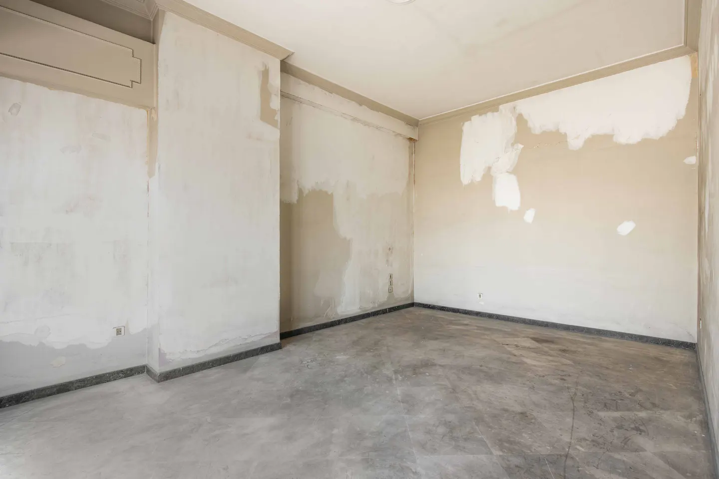 Empty room with unfinished walls and concrete floor. The walls are beige with white patches. Dark trim runs along the base.