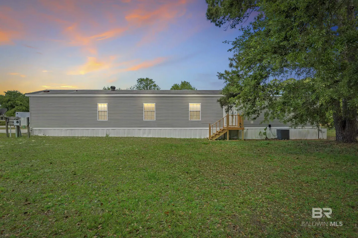 Exterior view of a gray, single-story home with a wooden porch and green lawn under a colorful sunset sky.