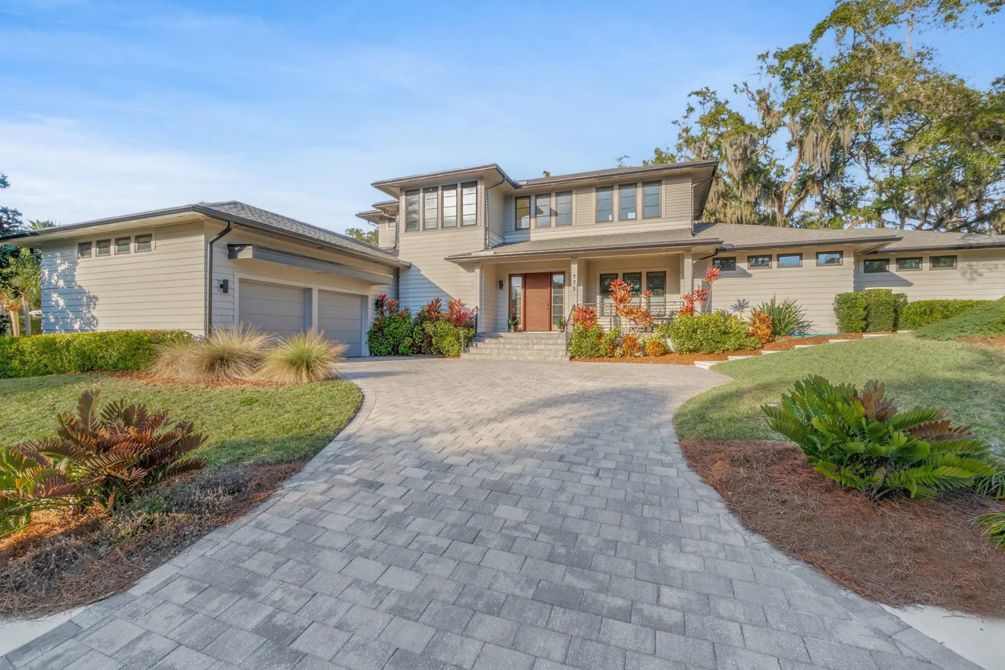 Two-story beige house with a gray brick driveway, green lawn, and red and green bushes.