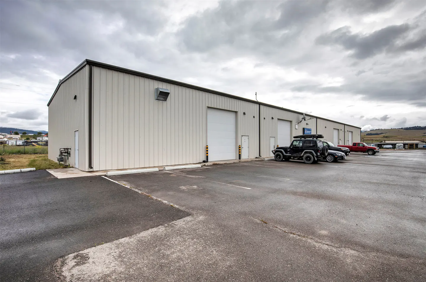 Exterior view of a beige metal industrial warehouse with multiple garage doors and a paved parking lot under a cloudy sky.