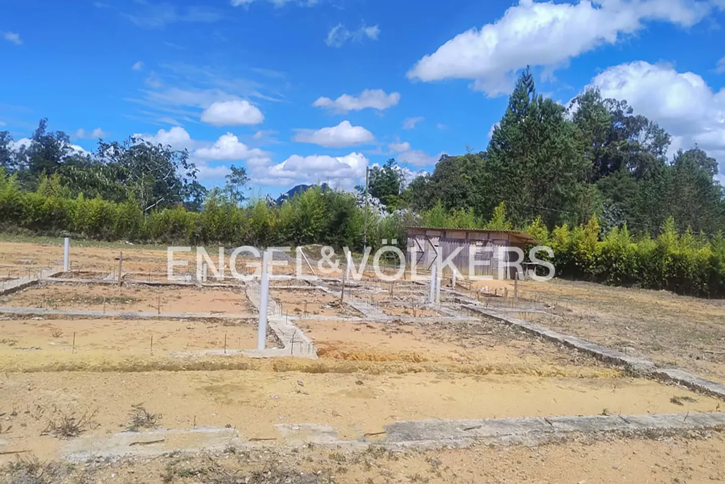 A vacant lot with a concrete foundation and Engel & Volkers logo, under a blue sky with scattered clouds.