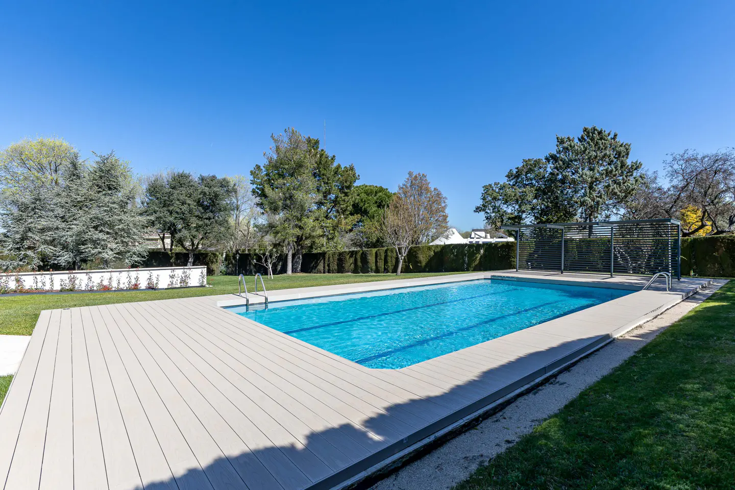 Outdoor pool with turquoise water, surrounded by a gray deck, green grass, and trees under a clear blue sky.
