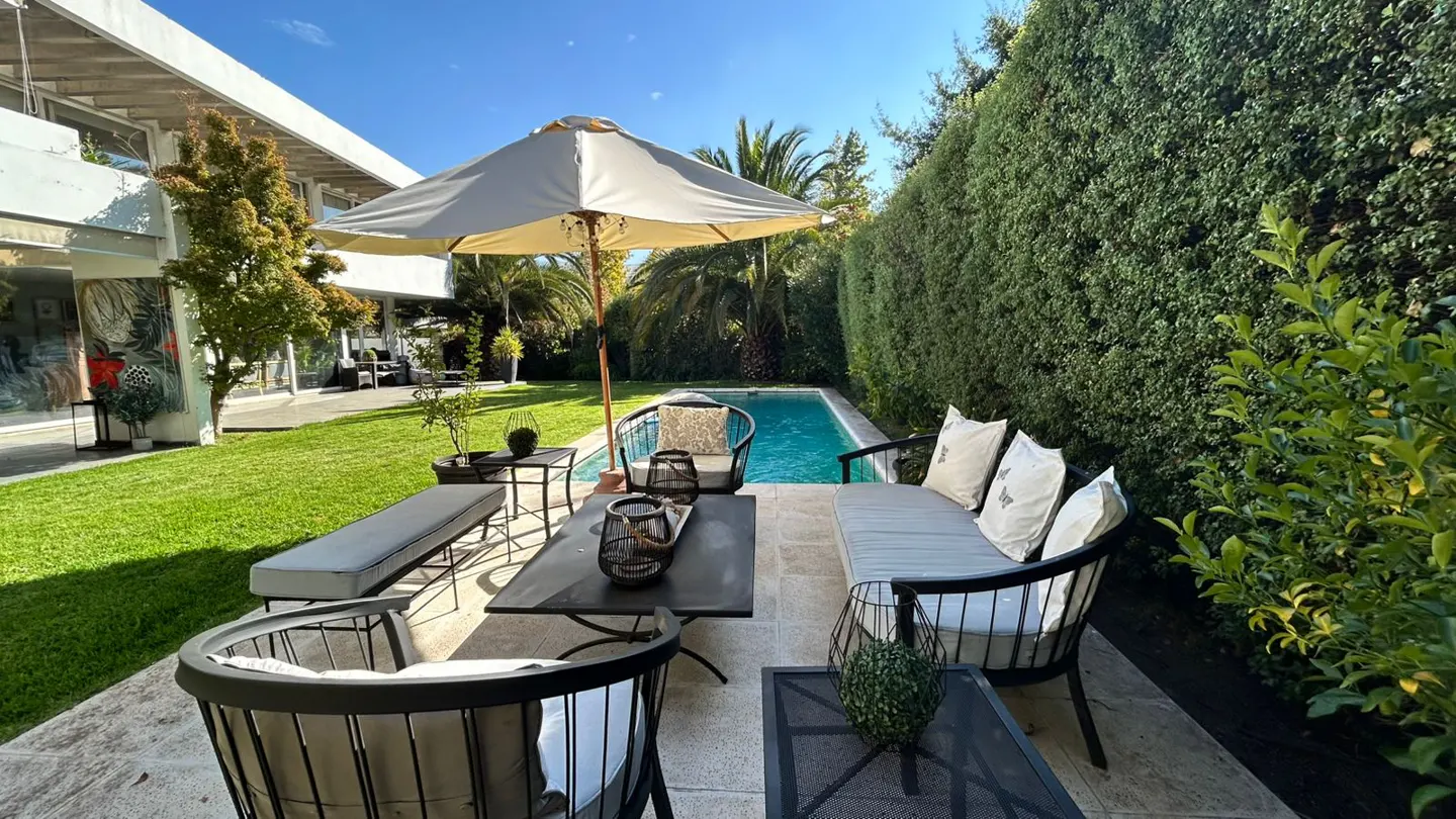 Outdoor patio with black furniture, a beige umbrella, and a pool surrounded by green hedges and grass.