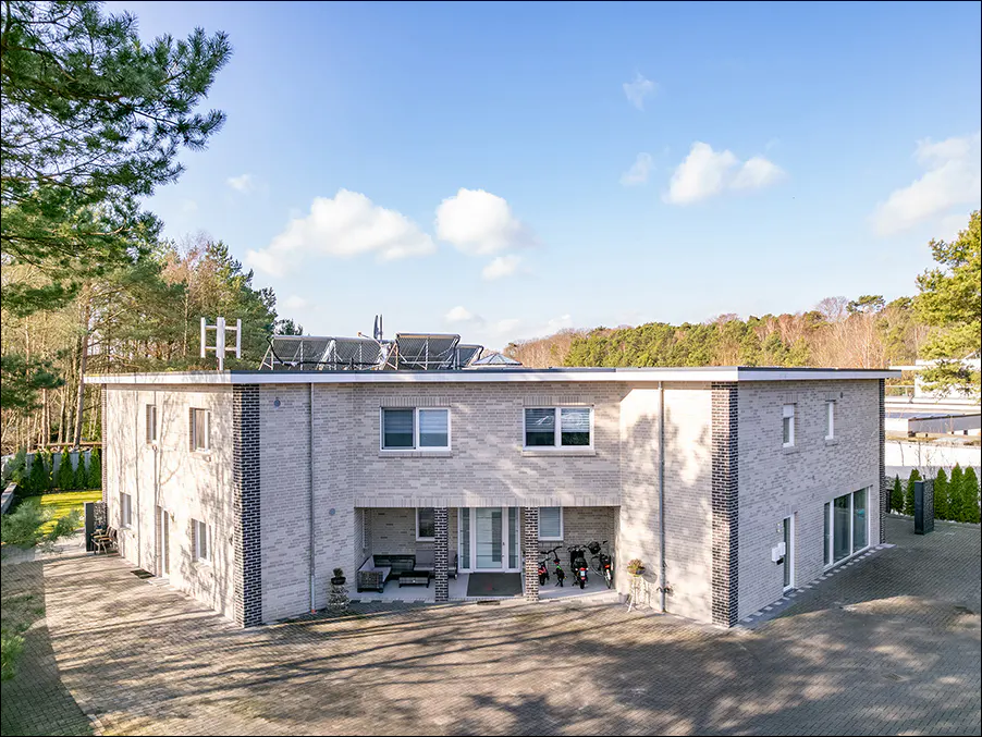 Two-story grey brick house with solar panels on the roof. A paved driveway leads to the entrance. Trees surround the property.