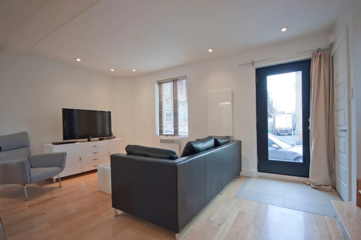 Modern living room with light wood floors, a black leather sofa, grey armchair, TV on a white console, and a glass door.