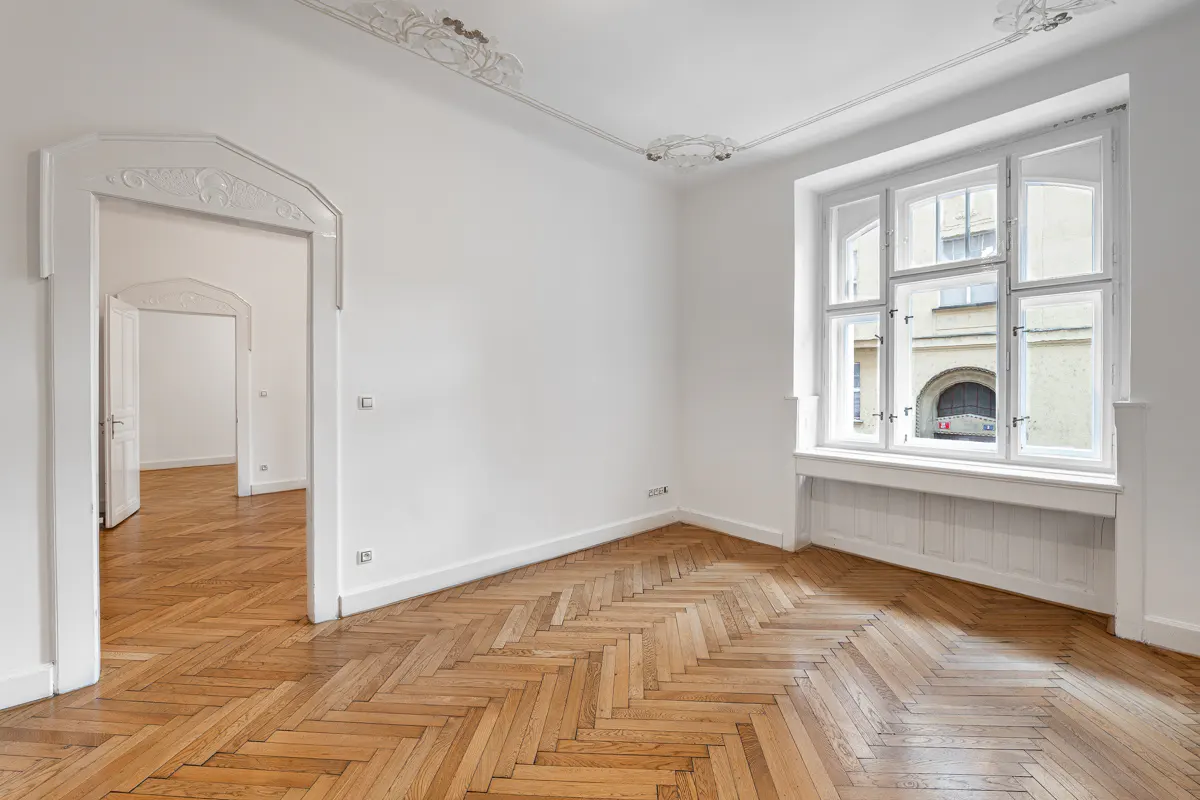 Empty white room with herringbone wood floors, large window, and ornate doorway leading to another room.