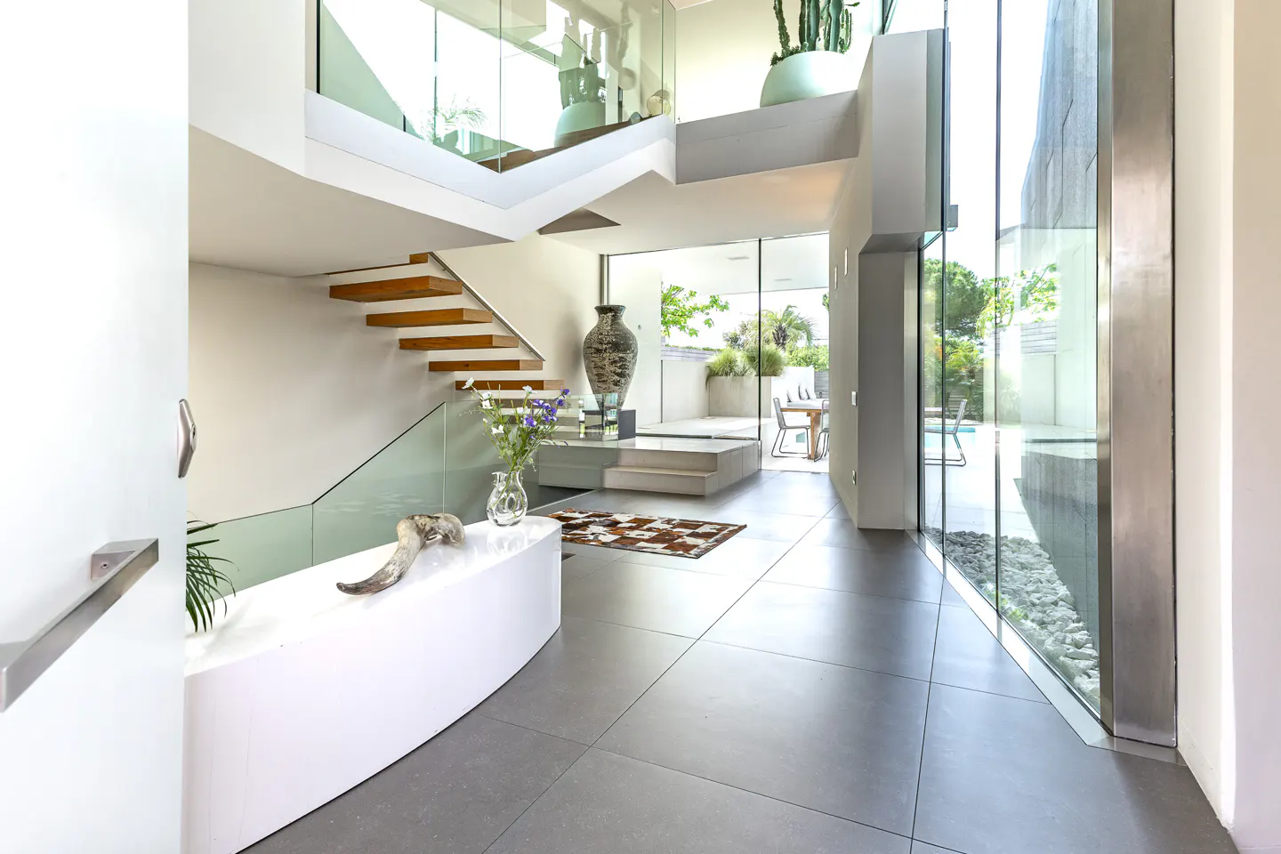 Modern home foyer with gray tile floors, white walls, and wood stairs. Glass railings and large windows provide natural light.