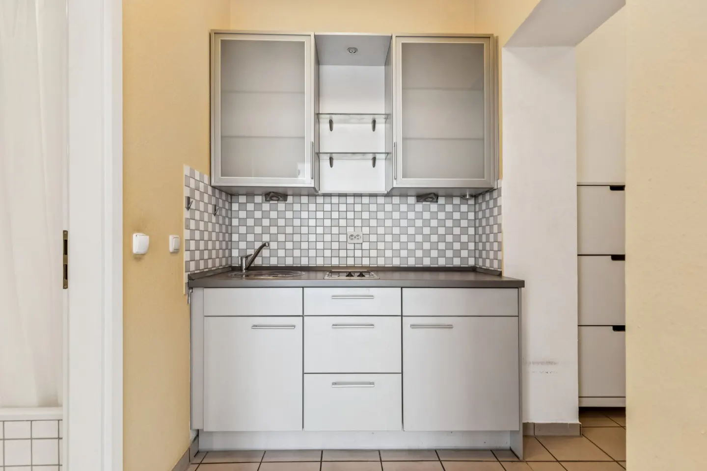 A small kitchen with white cabinets, a gray countertop, and a checkered tile backsplash. There are frosted glass cabinets above the counter.