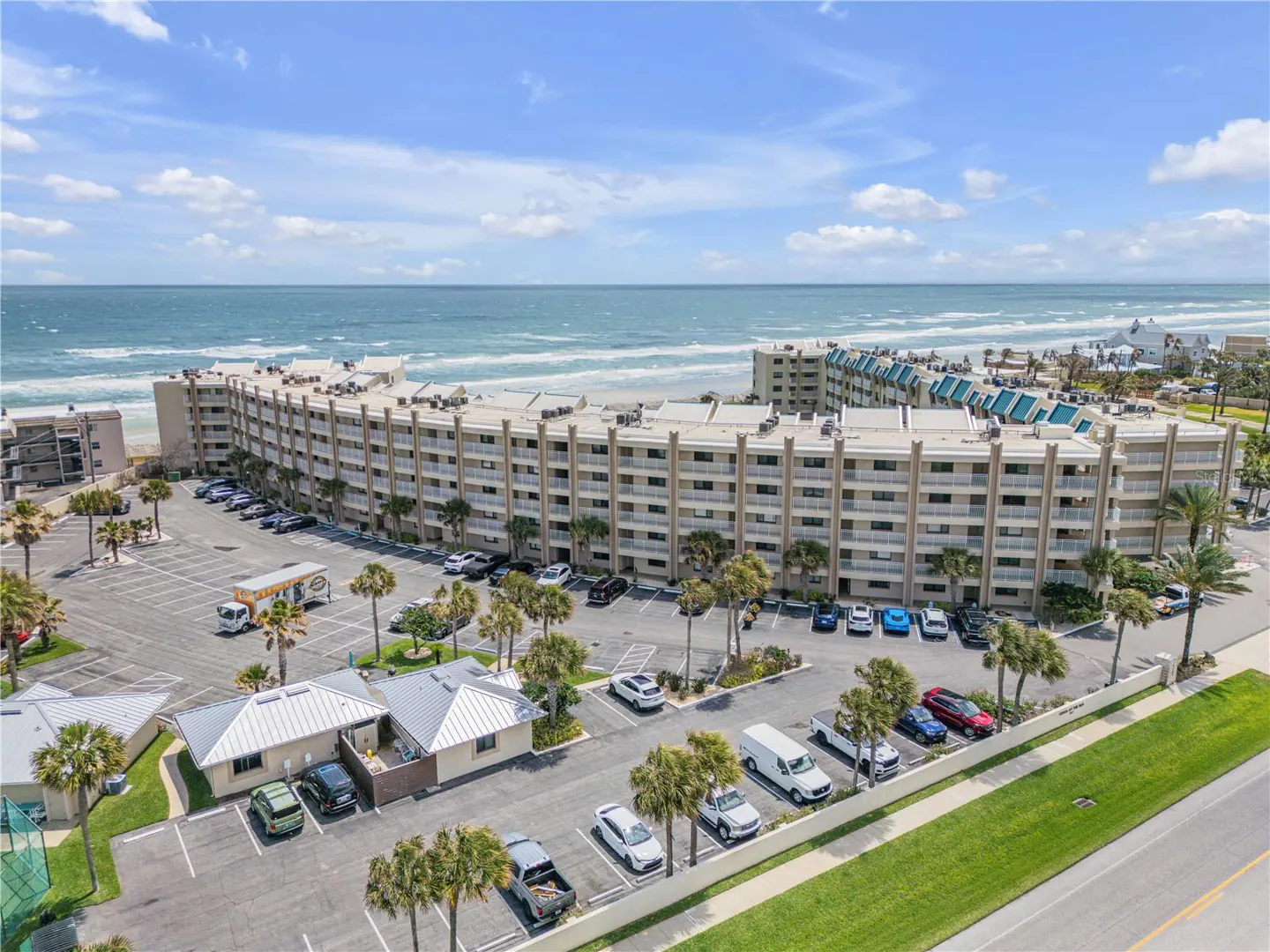 Aerial view of a beige condo building with balconies, a parking lot, palm trees, and the ocean in the background.