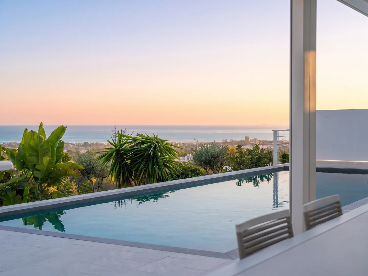 Infinity pool overlooking the ocean at sunset. Palm trees and greenery frame the view. White chairs are in the foreground.