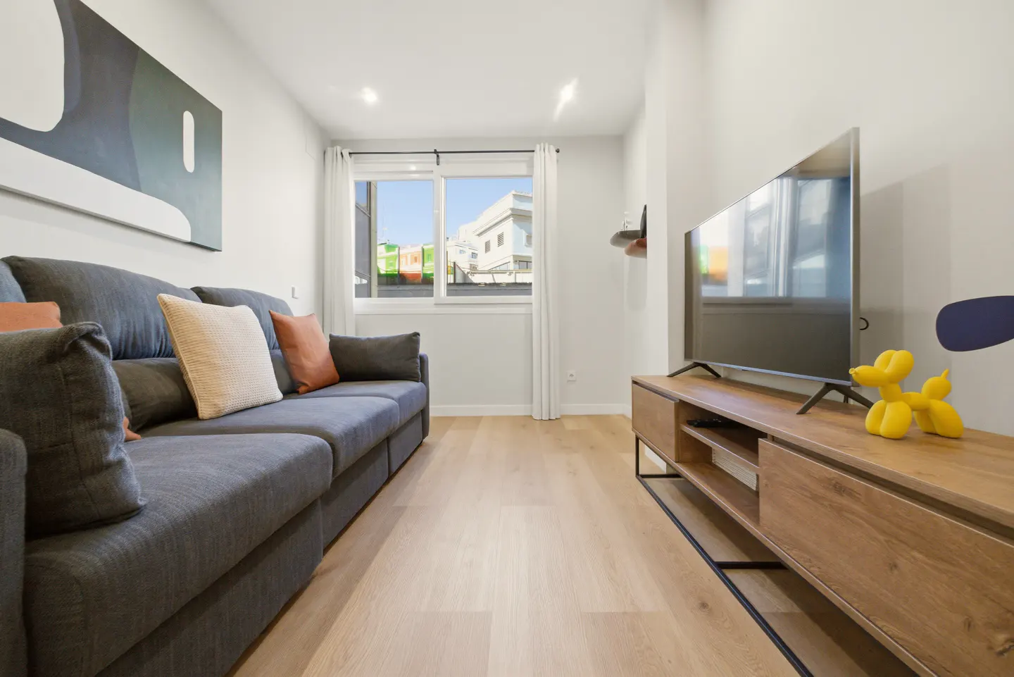 Living room with gray sofa, wood floors, and a TV on a wood stand with a yellow balloon dog. A window shows a building outside.